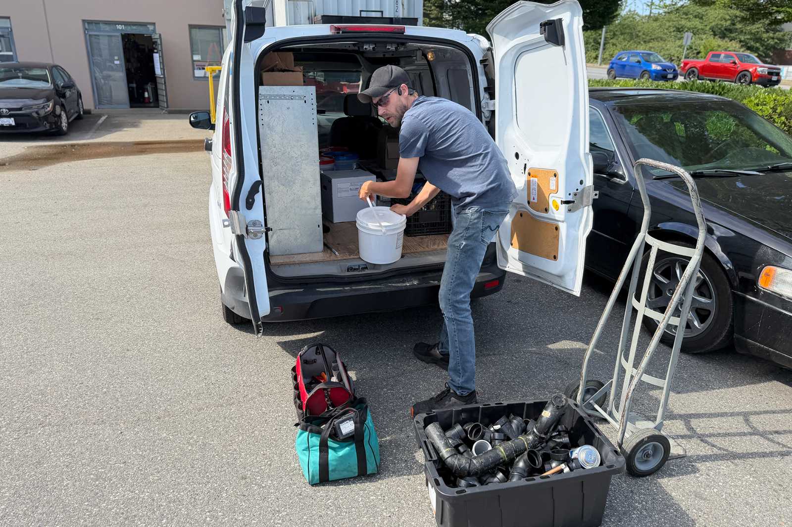 Commercial dishwasher technician gathering plumbing supplies and tools from the back of a service van for an installation job.